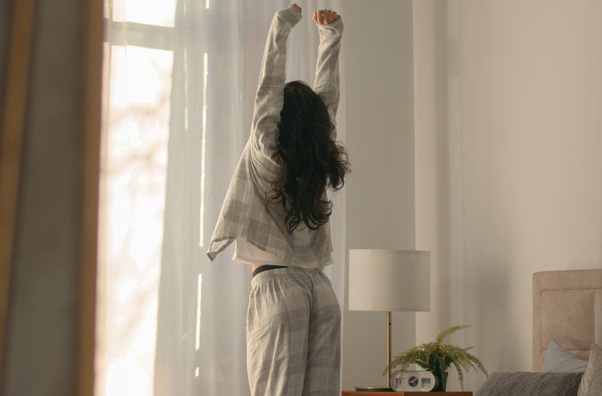 A woman is seen stretching gracefully in front of a window, with sunlight, refreshed after a good nights sleep